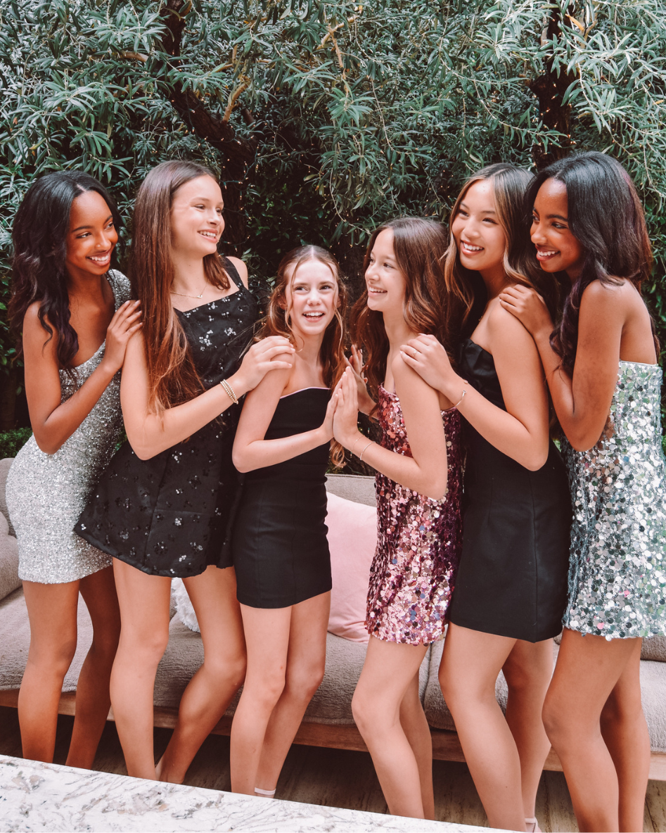 Group of six girls in fancy dresses posing together outdoors with greenery in the background