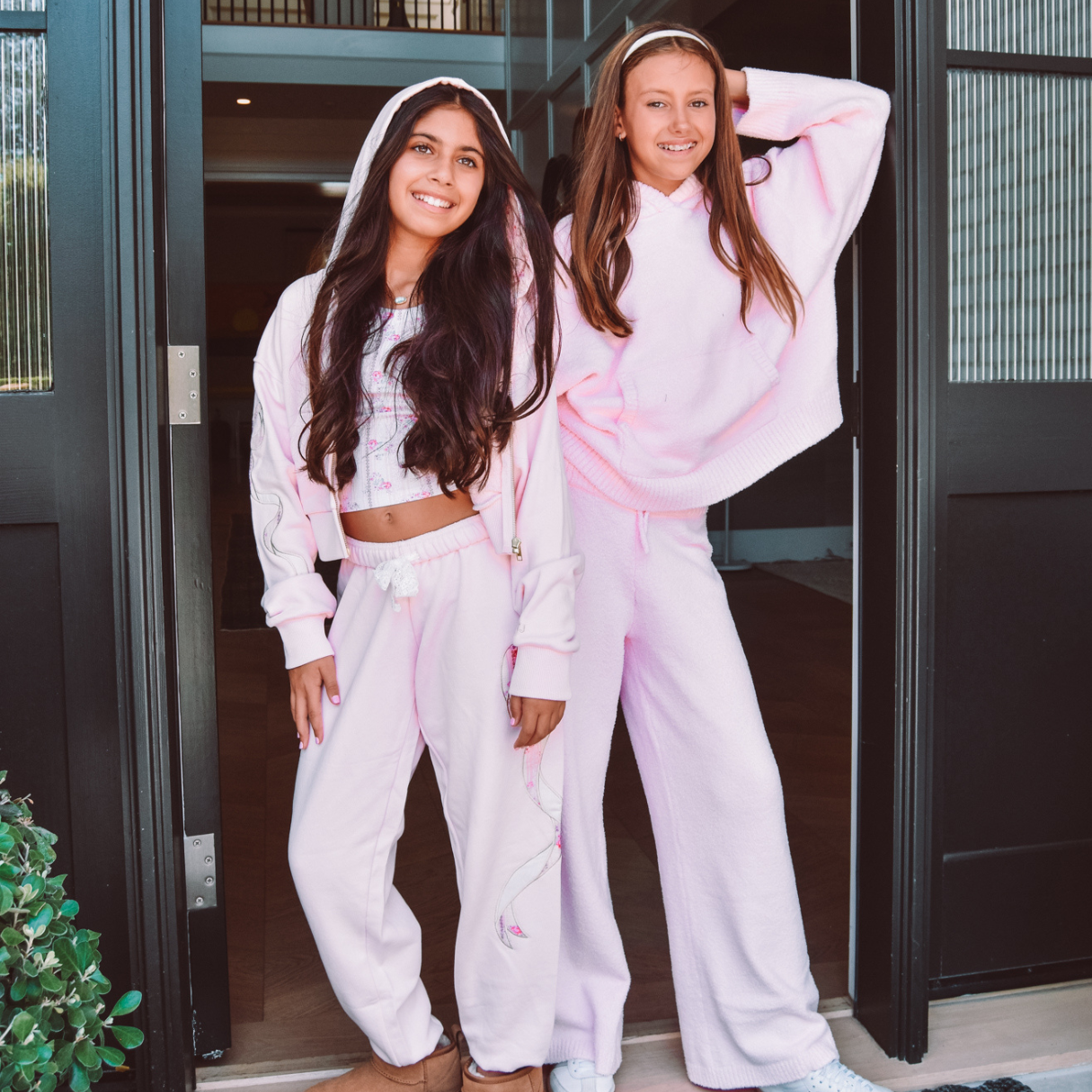 Two tweens in cozy pink loungewear pose in front of an open door.