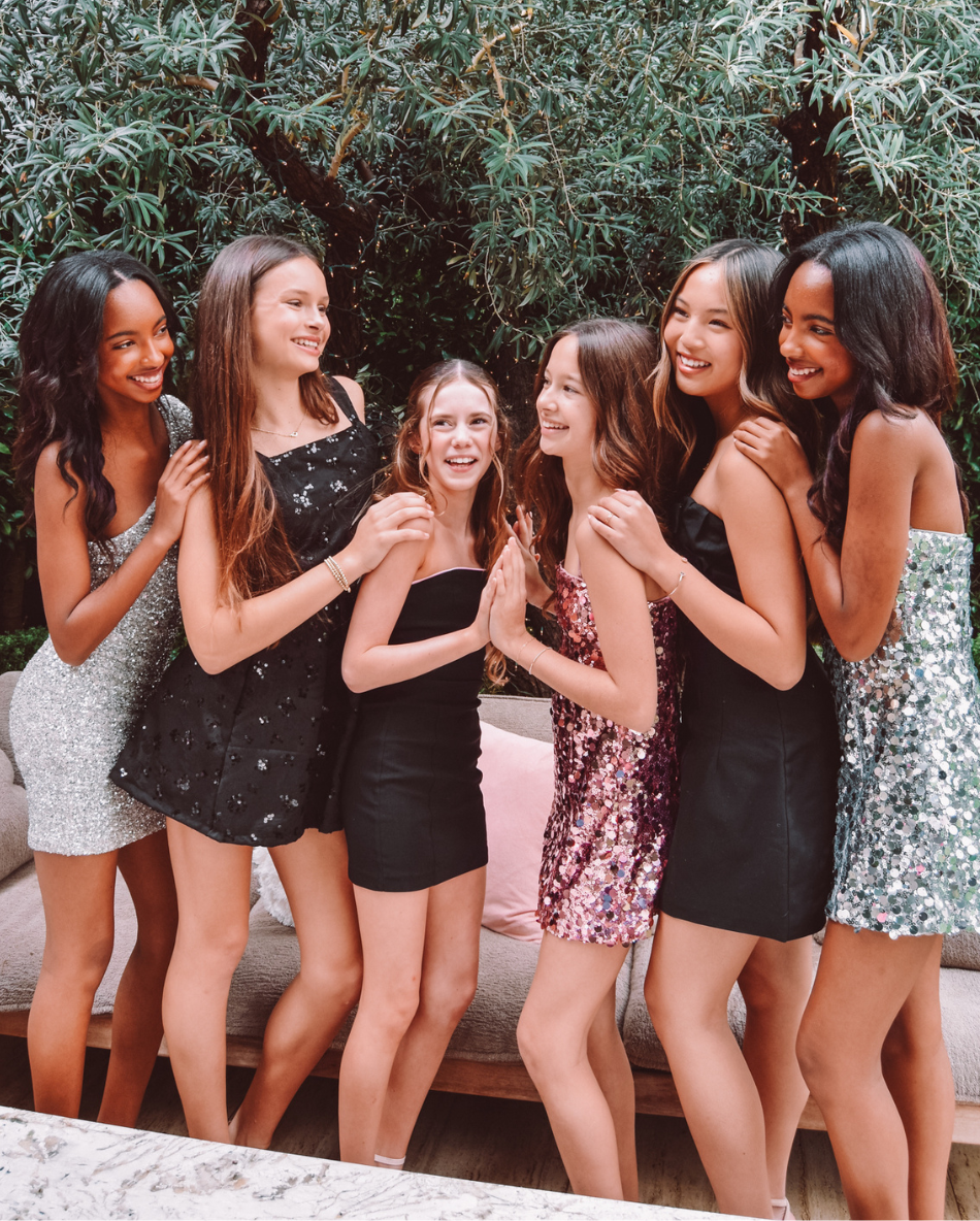 Group of six girls in fancy dresses posing together outdoors with greenery in the background
