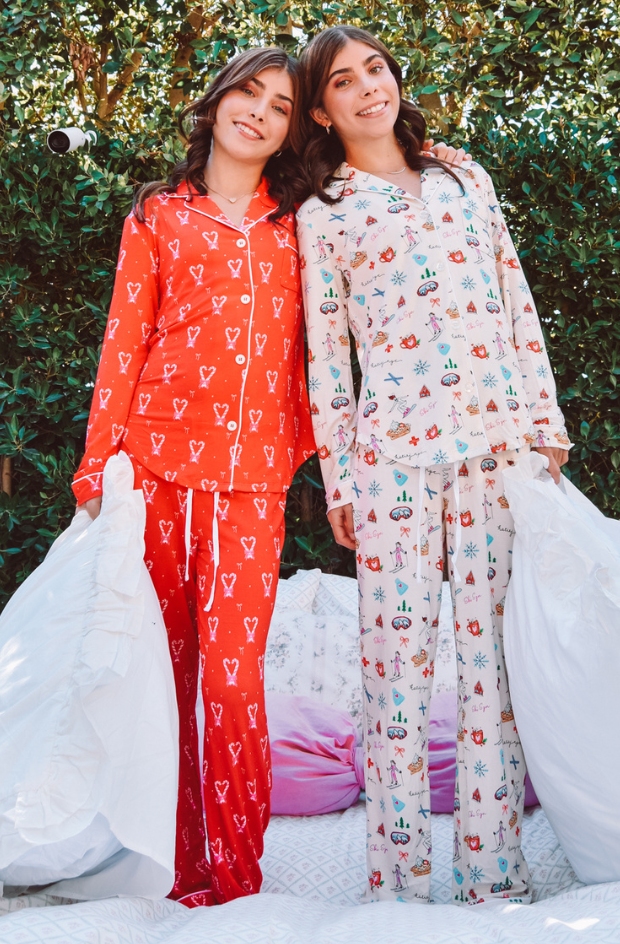 Identical twins with dark hair stand in holiday pajamas holding pillows.