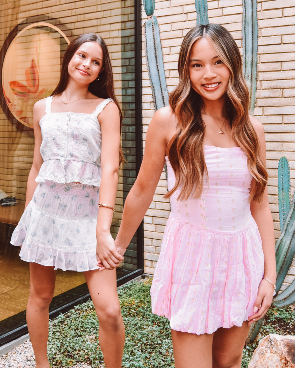 Two girls holding hands in front of a brick wall with cacti. They are dressed in pastel sundresses.