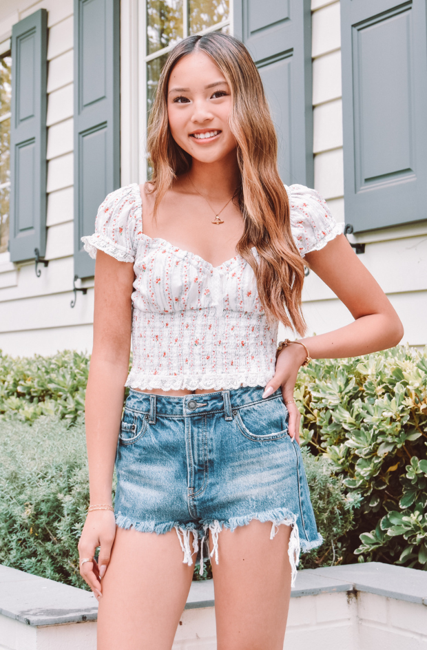 Young woman wearing a white floral top and denim shorts standing in front of a house.