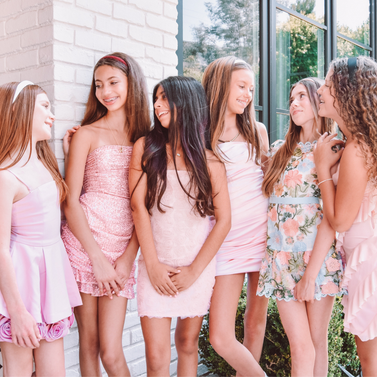 Six young women standing together outdoors in light pink and floral dresses, posing and smiling.