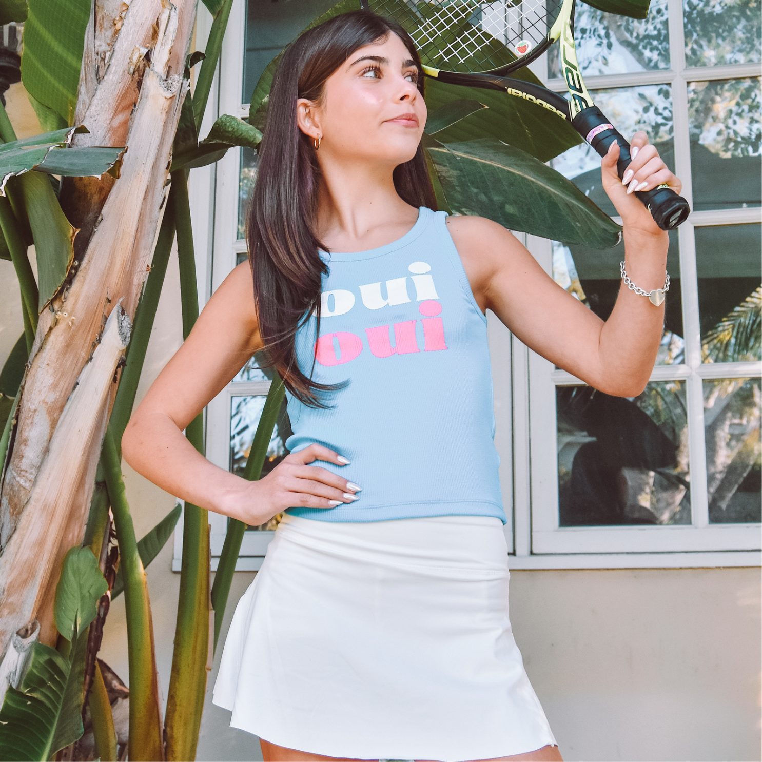 A girl with dark hair stands with her arm up holding a tennis racket. She is wearing a white athletic skirt and a blue tank top that says "Oui Oui"