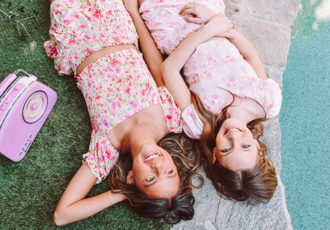 Two girls laughing while laying on the backs next to a pool and vintage radio. They are both wearing floral skirt and top matching sets.