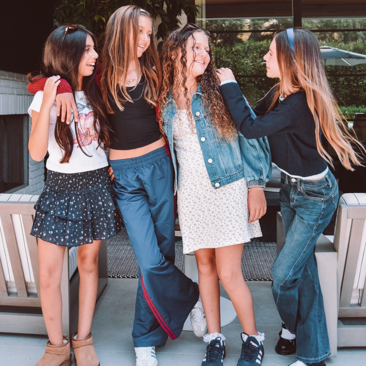 Four young women standing together on a patio, wearing casual, trendy outfits including a denim jacket, white dress, blue flared track pants, and jeans.